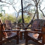 Rustic wooden chairs surround a small table on a stone patio at Moholoholo Forest Camp, offering a tranquil nature retreat